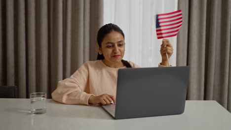 Happy-brunette-girl-with-Brown-skin-color-studies-foreign-languages-at-home-using-her-laptop-and-communicates-via-video-conference-while-holding-the-flag-of-the-United-States-of-America-at-home-in-a-modern-apartment