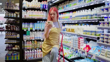 Happy-girl-with-pink-hair-supermarket-worker-takes-a-selfie-using-a-white-smartphone-during-a-break-from-work-in-the-dairy-department-in-the-supermarket