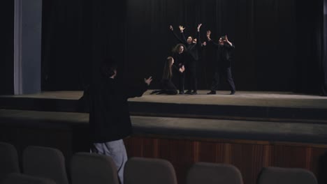 From-behind,-a-confident-male-director-holds-papers-and-gestures,-demonstrating-correct-acting-to-actors-in-black-suits-during-a-theater-stage-rehearsal