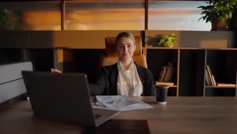 Portrait-of-a-confident-blonde-girl-in-a-business-uniform-who-sits-at-a-wooden-table-in-the-office-in-front-of-important-papers-and-a-laptop-in-a-sunny-office