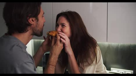 A-happy-brunette-guy-with-stubble-together-with-his-brunette-girlfriend-in-a-beige-T-shirt-simultaneously-bite-a-croissant-from-different-sides-during-a-romantic-date-in-the-kitchen-in-the-evening