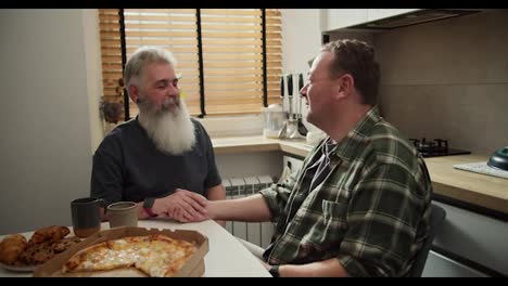 Happy-couple-of-LGBT-men-talking-and-holding-hands-while-sitting-in-the-kitchen-during-their-lunch.-An-elderly-man-with-gray-hair-and-a-lush-beard-in-a-gray-T-shirt-holds-hands-with-his-brunette-boyfriend-in-a-checkered-green-shirt-while-sitting-in-the-kitchen-at-the-table