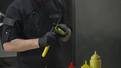 Close-up-of-a-professional-chef-a-man-in-a-black-uniform-and-wearing-protective-rubber-gloves-peels-an-avocado-before-starting-to-prepare-a-salad-in-a-restaurant-kitchen