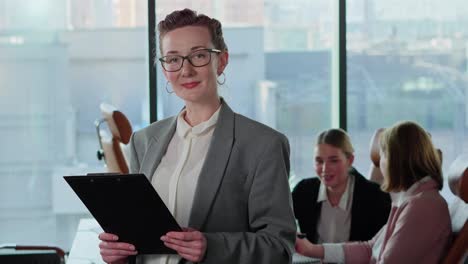 Retrato-De-Una-Chica-Rubia-De-Mediana-Edad-Segura-De-Sí-Misma-Con-Gafas-Y-Ropa-De-Trabajo-Sosteniendo-Una-Tableta-En-Sus-Manos-Cerca-De-Sus-Colegas-Empresarias-En-Una-Oficina-Moderna-Con-Grandes-Ventanales