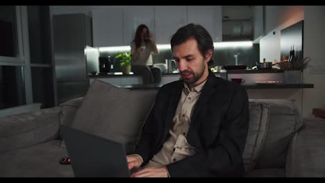 A-confident-brunette-man-with-stubble-in-a-black-jacket-and-beige-shorts-sits-on-the-sofa-and-works-at-a-gray-laptop-while-his-brunette-girlfriend-in-a-beige-T-shirt-sits-in-the-kitchen-and-drinks-tea