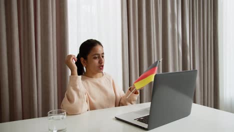 A-happy-brunette-girl-in-a-light-jacket-communicates-online-using-a-gray-laptop-while-studying-a-foreign-language-German-and-shows-a-flag-in-a-video-conference-at-home-online