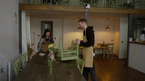 A-happy-blond-guy-in-light-brown-together-with-his-colleague-a-girl-waitress-lift-chairs-put-them-on-tables-and-clear-the-hall-in-a-cafe-after-a-working-day