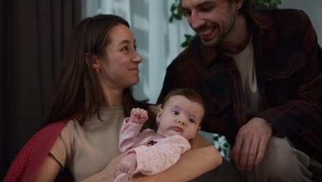 Happy-young-brunette-mother-in-a-red-sweater-holds-her-little-baby-daughter-in-her-arms-and-strokes-her-together-with-her-brunette-husband-in-a-modern-apartment