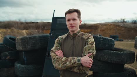 Portrait-of-a-confident-military-man-in-a-camouflage-green-uniform-who-folds-his-arms-on-his-chest-and-poses-with-a-serious-expression-near-barricades-made-of-car-tires-in-the-steppe-during-combat-training