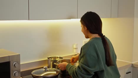 From-behind-a-happy-brunette-girl-in-a-green-jacket-and-a-black-apron-prepares-food-in-a-saucepan-in-a-modern-kitchen.-Happy-girl-in-apron-preparing-food-in-the-kitchen