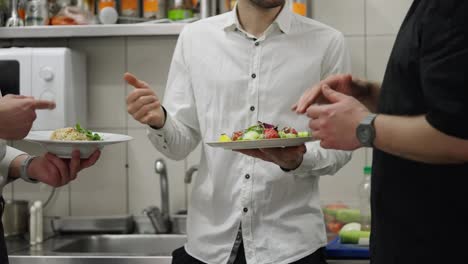 Close-up-of-a-cook-and-two-waiters-communicating-about-dishes-while-working-in-the-kitchen-in-a-restaurant.-Two-waiters-in-shirts-hold-salads-in-their-hands-and-communicate-with-the-chef-about-their-quality-in-the-kitchen