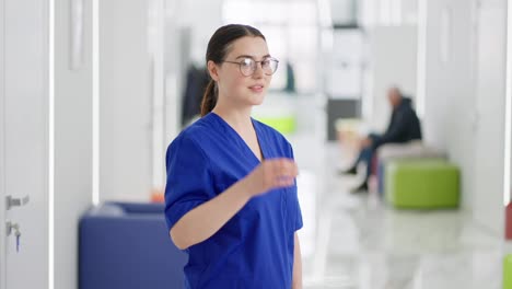 Portrait-of-a-confident-girl-brunette-doctor-in-round-glasses-and-a-blue-uniform-turns-her-head-and-poses-in-the-corridor-of-a-bright-clinic.-Girl-brunette-doctor-adjusts-her-glasses-and-confidently-poses-in-the-corridor-of-a-modern-clinic