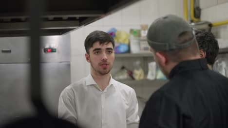 Por-Encima-Del-Hombro,-Un-Camarero-Con-Camisa-Blanca-Se-Comunica-Con-Un-Chef-Profesional-Con-Uniforme-Negro-Sobre-El-Plato-Y-La-Relación-Entre-Camareros-Y-Cocineros-En-La-Cocina-De-Un-Restaurante.