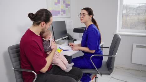 Confident-girl-doctor-in-glasses-and-blue-uniform-communicates-with-young-father-of-little-baby-girl-and-plays-with-baby-in-modern-clinic