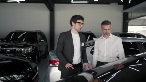 A-confident-middle-aged-man-with-a-gray-beard-and-a-white-shirt-communicates-with-a-young-brunette-guy-an-assistant-in-a-business-suit-in-a-car-dealership-and-examines-a-black-car