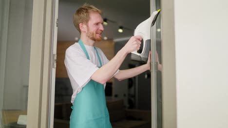 Side-view-of-a-confident-male-blond-cleaner-in-a-white-T-shirt-and-a-gray-blue-apron-washes-the-glass-in-the-door-using-a-vacuum-cleaner-for-windows-in-a-modern-apartment-while-calling-a-cleaning-company