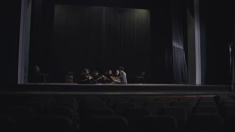 View-from-afar-a-group-of-confident-young-actors-in-black-suits-sit-on-stage-in-a-theater-and-have-lunch-during-a-break-during-a-rehearsal-in-a-large-theater-with-a-black-curtain