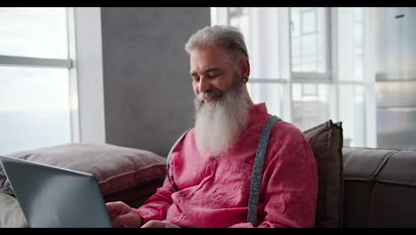 A-serious-and-happy-elderly-man-with-gray-hair-and-a-lush-beard-in-a-pink-shirt-sits-on-a-sofa-in-a-modern-apartment-and-works-on-a-laptop