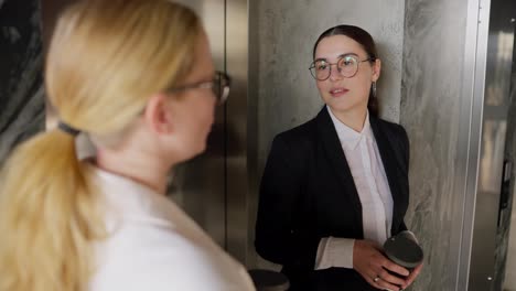 Over-the-shoulder-A-confident-brunette-girl-in-round-glasses-and-a-business-suit-communicates-with-her-middle-aged-blonde-colleague-during-a-short-break-and-drinking-coffee-in-a-modern-office