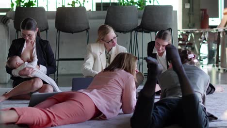 Side-view-of-a-group-of-confident-businesswomen-in-business-clothes-sitting-on-a-soft-carpet-in-the-office-and-communicating-with-each-other-while-their-colleague-a-brunette-girl-in-business-clothes-breastfeeds-her-infant-child-in-the-office