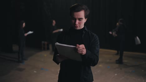 Portrait-of-a-confident-and-happy-brunette-male-actor-in-a-black-suit-who-reads-his-text-and-then-folds-piles-of-papers-near-his-chest-near-the-actors-who-are-preparing-to-perform-on-stage-in-the-theater