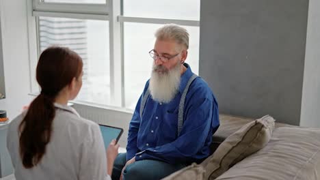 An-elderly-Serbian-man-with-hair-and-a-white-lush-beard-in-glasses-and-a-blue-shirt-communicates-with-a-brunette-girl-doctor-in-a-medical-gown-who-is-holding-a-tablet-in-her-hands-and-recording-the-patient’s-complaints-during-a-home-examination