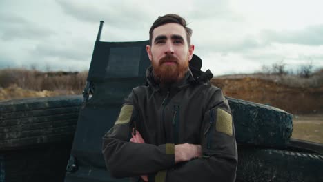 Portrait-of-a-confident-brunette-man-with-a-beard-in-dark-green-army-clothes-near-car-tires-in-the-steppe-during-combat-training.-Confident-male-military-man-posing-during-combat-exercise-outside-the-city