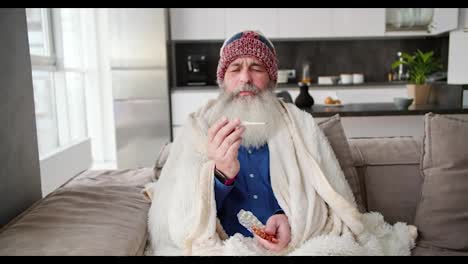 Portrait-of-an-elderly-man-in-a-red-cap-and-a-white-blanket-sits-and-checks-his-temperature-with-a-thermometer-and-also-holds-a-set-of-pills-in-his-hand-while-sitting-on-a-brown-sofa-in-a-modern-apartment