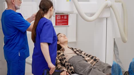 A-young-brunette-girl-in-a-checkered-shirt-lies-down-on-the-bed-during-her-fluorography-under-the-supervision-of-an-experienced-male-and-female-doctor-in-a-modern-clinic