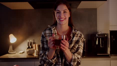 Portrait-of-a-happy-and-cheerful-brunette-girl-in-a-checkered-shirt-who-blows-on-a-cup-of-hot-tea-in-her-hands-in-a-modern-apartment-in-the-kitchen-in-the-evening