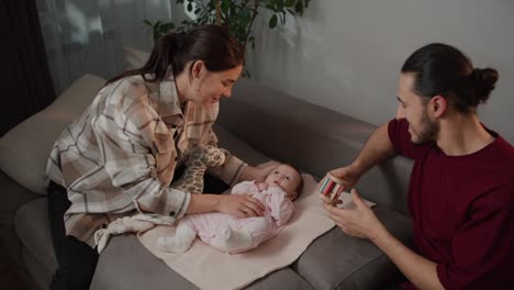 Happy-brunette-girl-in-a-plaid-shirt-together-with-her-brunette-husband-in-a-red-T-shirt-play-with-their-little-daughter-using-a-variety-of-toys-for-children-as-a-baby-in-a-pink-overalls-sitting-on-a-gray-sofa-in-a-modern-apartment