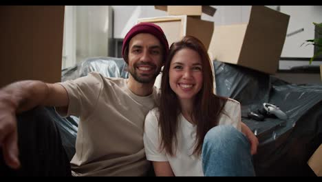 Close-up-portrait-of-a-happy-brunette-man-with-stubble-together-with-his-brunette-girlfriend-in-a-white-T-shirt-they-are-sitting-near-the-sofa-packed-in-a-black-plastic-case-among-a-large-number-of-boxes-in-a-new-apartment-after-moving