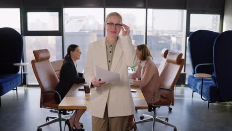 Portrait-of-a-confident-middle-aged-blonde-woman-who-takes-off-her-glasses-and-poses-in-a-white-business-suit-near-her-work-colleagues-In-the-office-at-a-table-in-an-office-with-large-windows