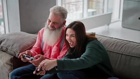 A-brunette-girl-in-a-green-sweater-plays-with-her-elderly-father-with-gray-hair-and-a-lush-beard-in-a-video-game-using-a-joystick-they-have-fun-and-rejoice-while-sitting-on-a-modern-sofa-in-an-apartment-with-large-windows-overlooking-the-sea