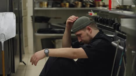 Close-up-of-a-tired-male-cook-in-a-black-uniform-takes-off-his-hat-and-straightens-his-hair-while-sitting-on-the-floor-leaning-on-the-table-in-the-kitchen-of-a-restaurant