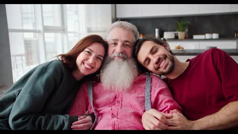 Portrait-of-a-Happy-man-with-gray-hair-and-a-lush-beard-in-a-pink-shirt-along-with-his-adult-children-a-brunette-man-and-a-girl-in-a-Green-jacket-in-a-modern-apartment