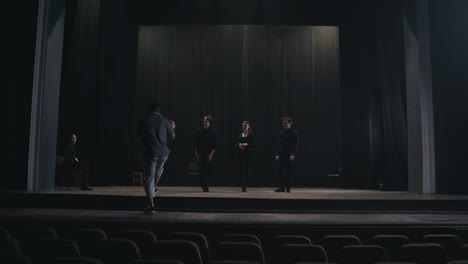 A-confident-stage-director-in-a-gray-T-shirt-comes-on-stage-to-explain-to-the-actors-how-to-play-and-move-correctly-on-stage-during-preparation-for-a-performance-in-an-empty-theater-with-black-curtains