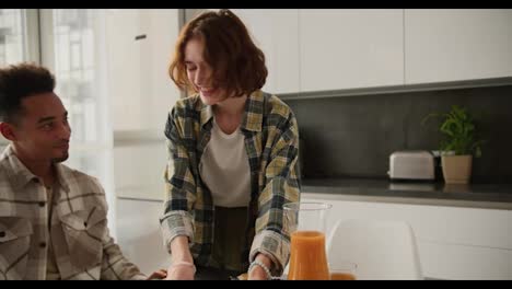 Happy-young-adult-girl-with-brown-hair-and-bob-hairstyle-serves-fresh-croissants-to-the-table-during-breakfast-with-her-young-Black-brunette-boyfriend-at-the-table-in-a-modern-apartment-in-the-kitchen