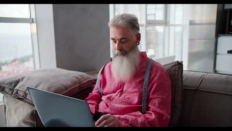 A-confident-and-happy-man-with-gray-hair-and-a-lush-beard-of-advanced-age-in-a-pink-shirt-sits-on-a-brown-sofa-and-types-on-his-gray-laptop-in-a-modern-apartment