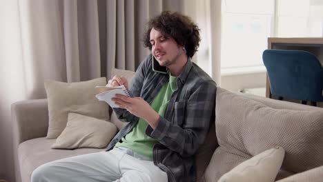 Confident-brunette-man-with-curly-hair-in-a-gray-checkered-shirt-communicates-on-the-phone-and-takes-notes-in-a-notepad-at-home-on-the-sofa