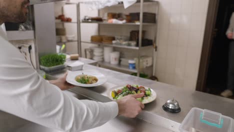 Un-Cocinero-Con-Camisa-Blanca-Coloca-Los-Platos-Preparados-Y-Las-Ensaladas-En-La-Mesa,-Avisa-Mediante-Una-Campana-Y-El-Camarero-Recoge-Los-Platos-Llevándolos-Al-Salón-Del-Restaurante.