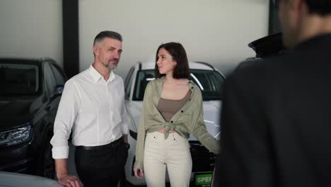 Una-Familia-Segura-De-Sí-Misma-En-Una-Sala-De-Exposición-De-Automóviles.-Un-Hombre-De-Mediana-Edad-Con-Una-Camisa-Blanca-Y-Su-Esposa-Morena-Se-Comunican-Emocionalmente-Con-Un-Asistente-Con-Un-Traje-De-Negocios-En-Una-Sala-De-Exposición-De-Automóviles-Sobre-Automóviles-Modernos.