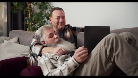 A-happy-middle-aged-man-with-gray-hair-and-a-lush-gray-beard-in-a-checkered-shirt-lies-on-the-lap-of-his-brunette-boyfriend-in-a-checkered-shirt-and-holds-a-gray-laptop-in-his-arms-during-their-common-pastime-near-the-laptop-on-the-sofa-in-the-apartment