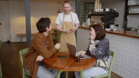 A-blond-guy-in-a-light-brown-apron-waiter-takes-an-order-from-a-couple-of-a-brunette-girl-and-a-guy-in-a-brown-jacket-in-a-cafe