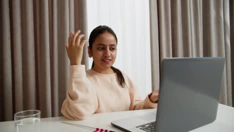 A-brunette-Indian-girl-in-a-light-jacket-studies-English-online-during-her-video-conference-and-shows-the-American-flag-to-the-camera-while-introducing-herself-in-a-modern-apartment-during-the-day