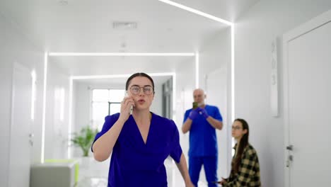 A-girl-doctor-with-glasses-and-a-blue-uniform-runs-down-the-corridor-talking-on-the-phone-and-runs-to-a-call-in-a-modern-clinic.-A-confident-girl-doctor-in-a-blue-uniform-hurries-on-call-and-runs-along-the-corridor-in-a-modern-bright-clinic