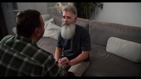 Over-the-shoulder-A-man-with-gray-hair-and-a-lush-gray-beard-in-a-gray-T-shirt-communicates-and-holds-the-hand-of-his-boyfriend-a-brunette-with-stubble-in-a-checkered-shirt-sitting-on-a-gray-sofa-in-the-evening-in-a-modern-apartment