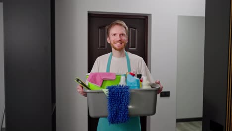 Portrait-of-a-confident-blond-professional-cleaner-man-in-a-blue-apron-who-holds-a-gray-plastic-basin-in-his-hands-which-is-filled-with-a-large-number-of-cleaning-tools-while-calling-a-cleaning-company-to-the-house-in-a-modern-apartment