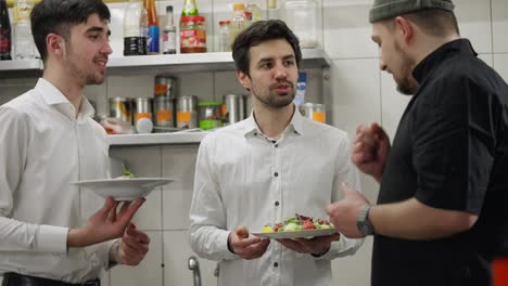 Los-Camareros-Confiados-Y-Alegres-Hablan-Con-El-Chef-Sobre-Las-Ensaladas-Que-Preparó-En-La-Cocina-De-Un-Restaurante.-Los-Chicos-Con-Camisas-Blancas-Hablan-Sobre-Ensaladas-Con-El-Chef-Que-Las-Preparó-En-La-Cocina-De-Un-Restaurante.