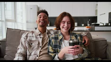 Portrait-of-a-happy-and-very-cheerful-couple-a-young-brunette-man-with-Black-skin-in-a-cream-plaid-shirt-sits-on-the-sofa-and-hugs-his-girlfriend-who-is-rejoicing-and-holding-a-mug-of-tea-in-her-hands-while-sitting-on-a-modern-sofa-in-a-modern-apartment